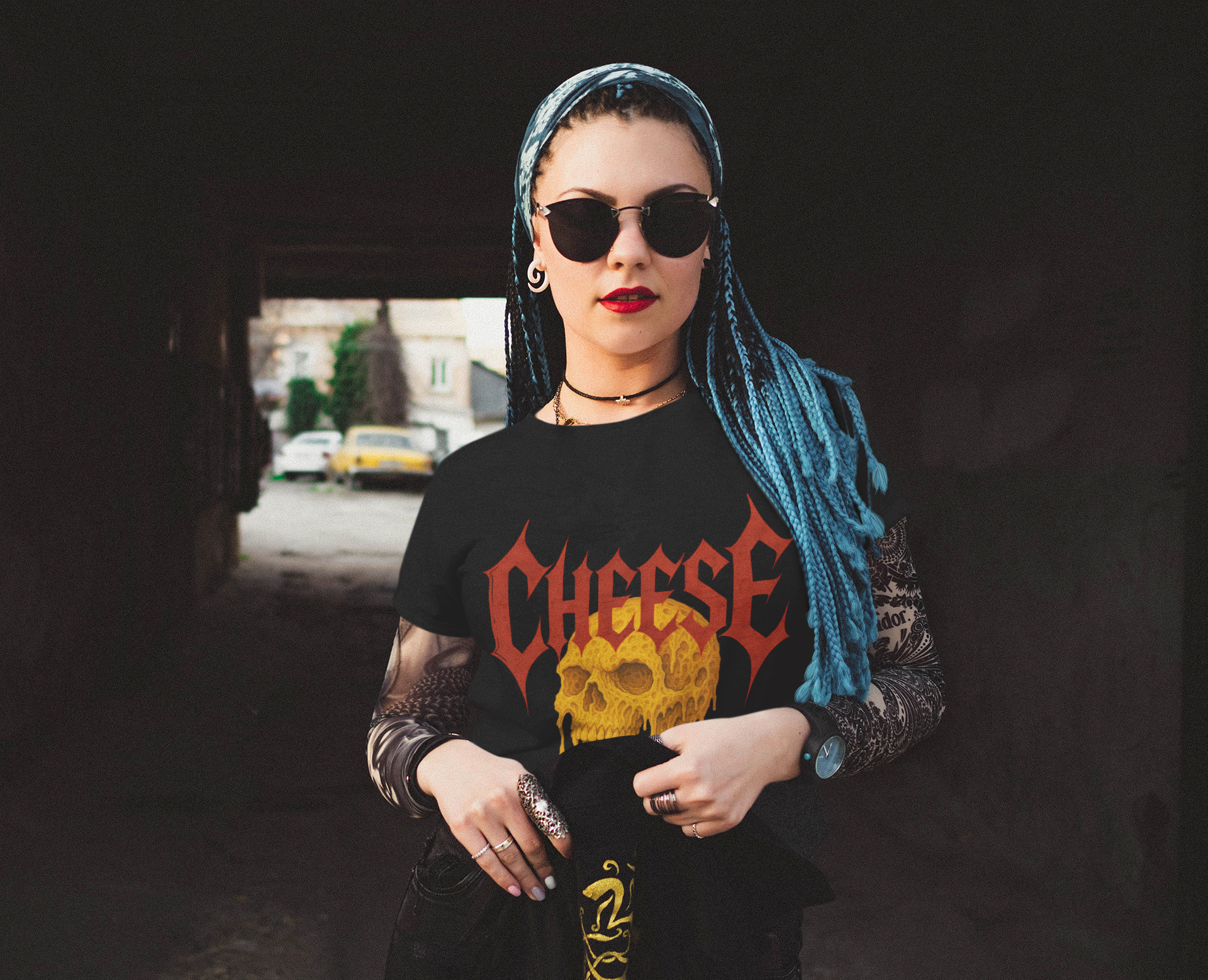 Person wearing a black t-shirt with 'CHEESE' and skull design, sunglasses, and blue hair in an urban setting.